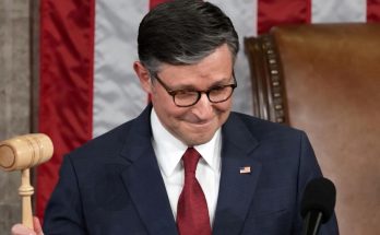A man in a dark suit and red tie holds a wooden gavel while standing at a podium with a