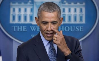 barack Obama at White House press briefing, touching nose, American flag and presidential