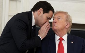 man in suit whispers secret to seated President Trump in office setting