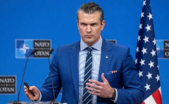 pete Hegseth stands at a podium at a NATO press conference, holding a pen, with an