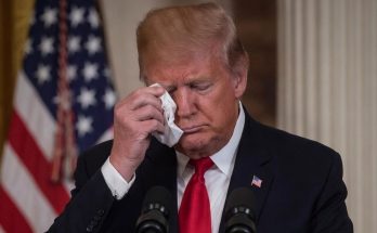 donald Trump wiping tear with tissue at podium with American flag behind him